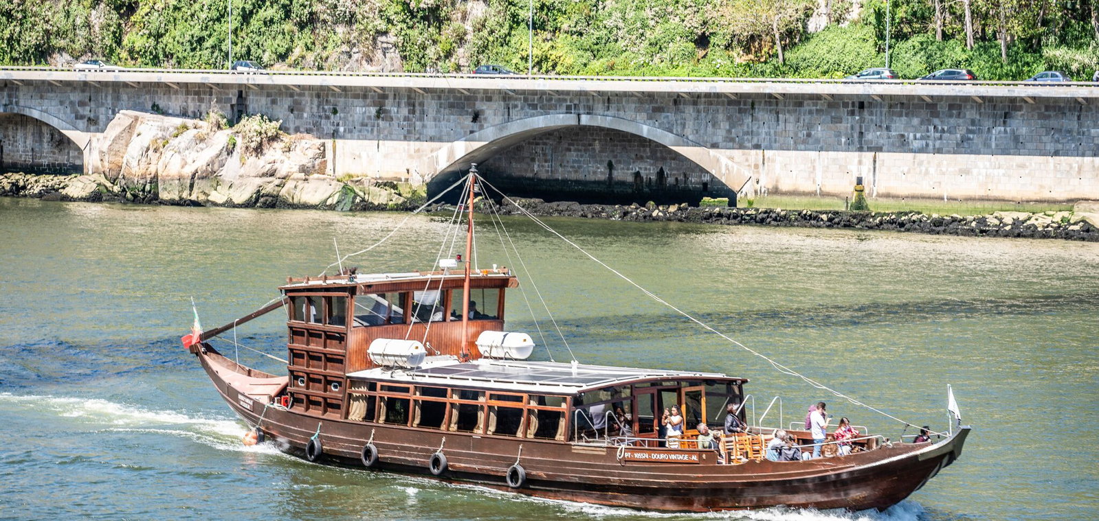A traditional wooden rabelo-style boat carrying passengers on the Douro beneath a stone bridge
